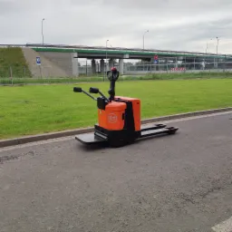 Pomarańczowo-czarny elektryczny wózek paletowy marki BT stoi na asfaltowej powierzchni, w tle widoczny wiadukt i zielony trawnik.