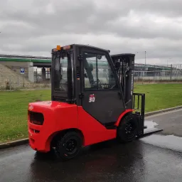 Czerwono-czarny wózek widłowy marki Hangcha, model 30, na zewnątrz na tle wiaduktu i zieleni, widok z boku na mokrej nawierzchni.