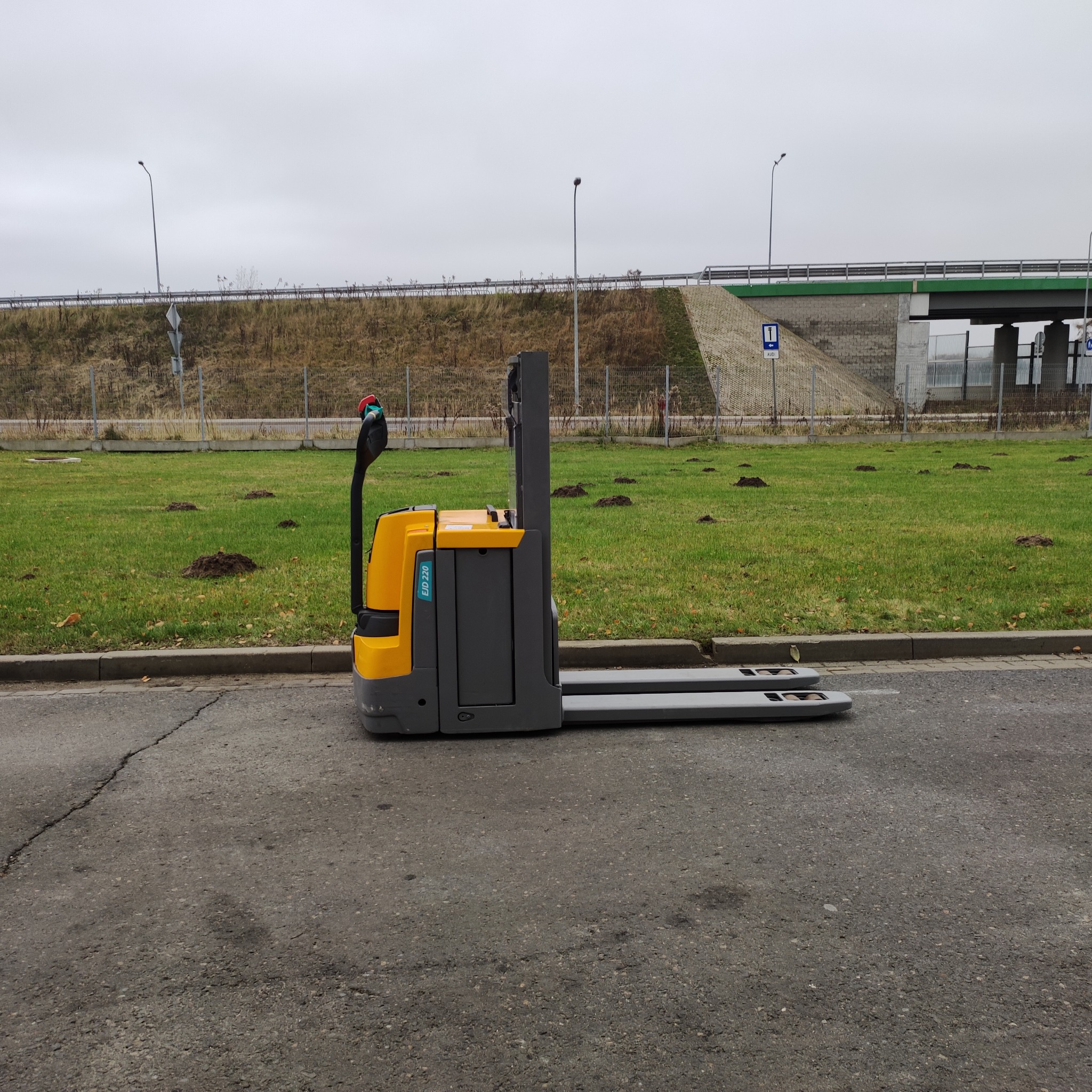 Żółto-szary wózek paletowy elektryczny EJD 220 stoi na asfalcie, w tle trawnik z kretowiskami i wiadukt autostradowy w pochmurny dzień.