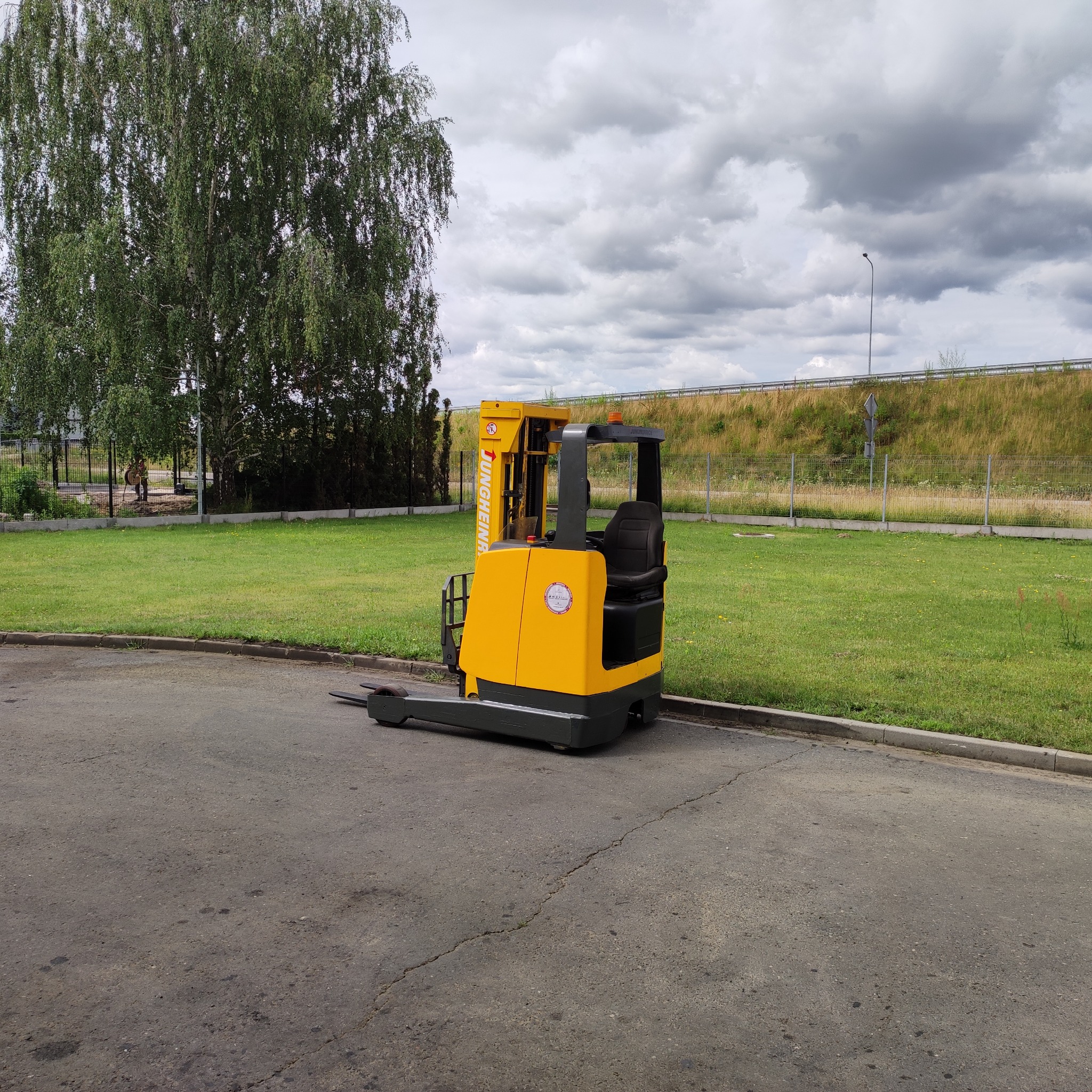 Żółto-czarny wózek paletowy elektryczny marki Jungheinrich, stojący na asfaltowej powierzchni, z trawnikiem i drzewami w tle pod pochmurnym niebem.