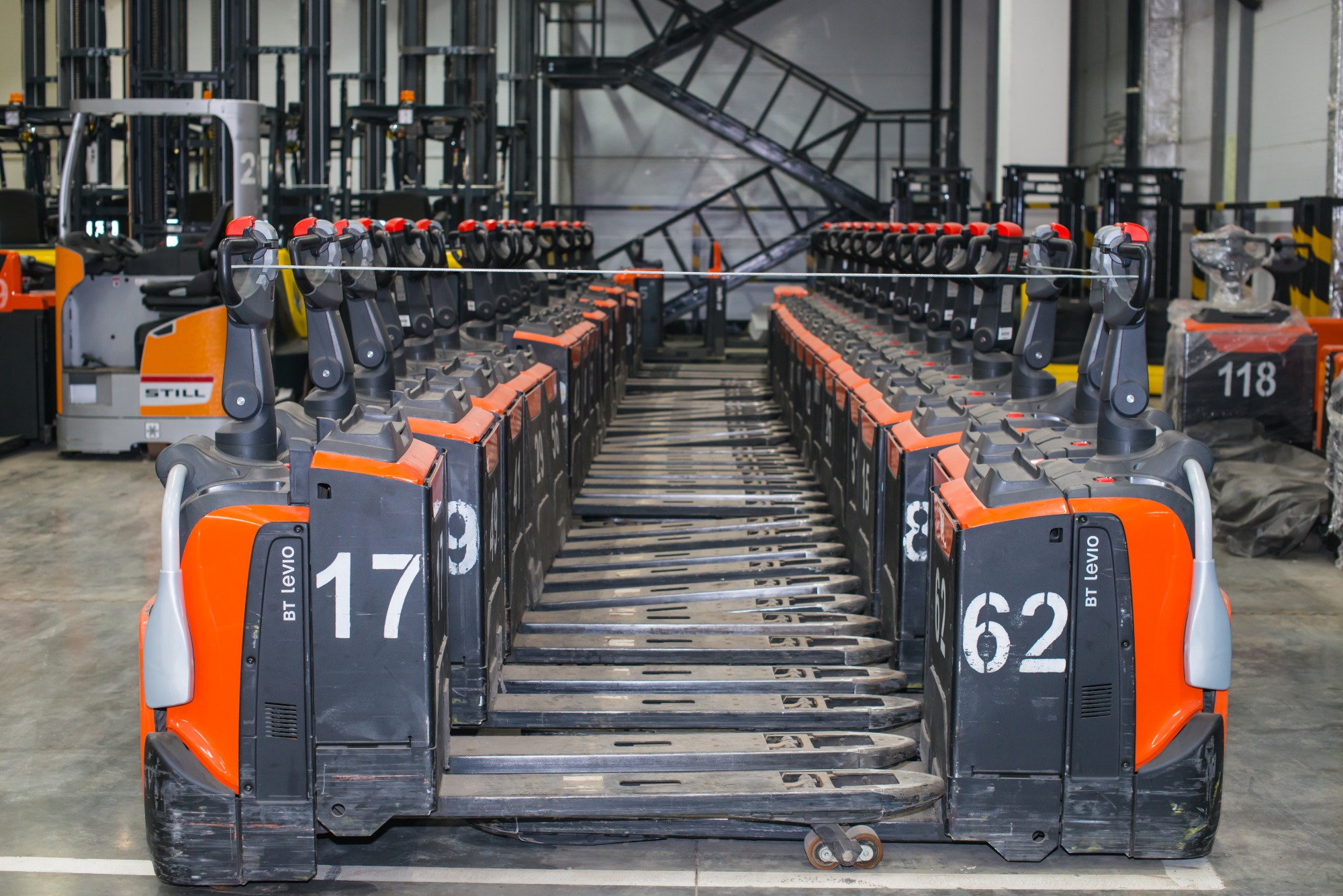Rząd zaparkowanych, pomarańczowo-czarnych wózków paletowych elektrycznych BT Levio z numerami 17 i 62 na boku, ustawionych w magazynie z widocznymi w tle wózkami widłowymi i metalową konstrukcją...