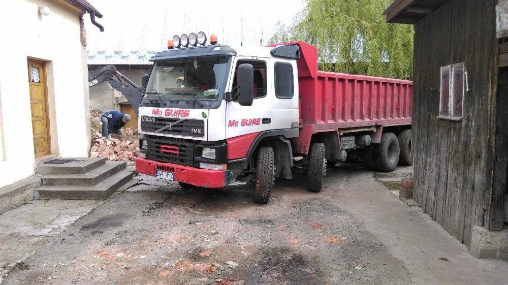 Ciężarówka wywrotka Volvo FM10 z czerwoną skrzynią ładunkową i napisem 'Mc Guire' na kabinie, zaparkowana na placu budowy z gruzem i koparką w tle.