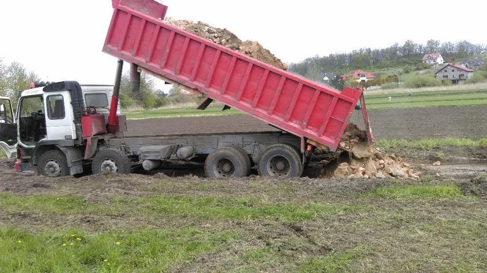 Ciężarówka wywrotka z podniesioną, czerwoną skrzynią ładunkową, wysypująca ziemię na błotniste pole, koła pojazdu częściowo zanurzone w grząskim terenie.