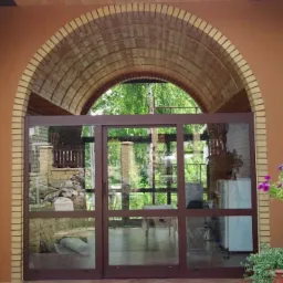 Zewnętrzne, przesuwne, przeszklone drzwi w brązowej ramie pod ceglanym łukiem, widok na taras i ogród.