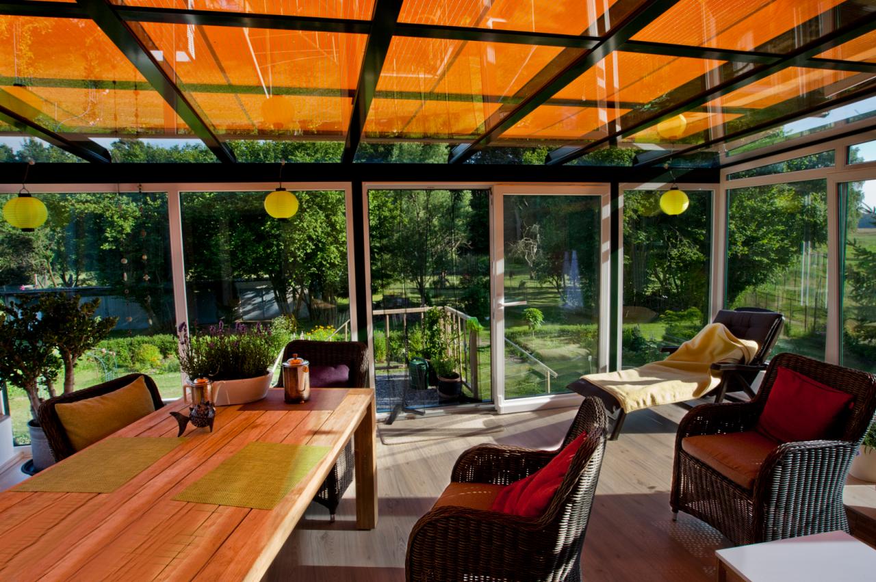 Jasna oranżeria z przeszklonym dachem i ścianami, drewnianym stołem, wiklinowymi fotelami i widokiem na zielony ogród z fontanną.