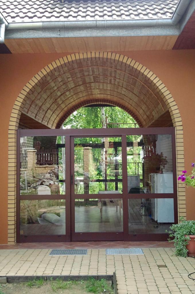 Zewnętrzne, przesuwne, przeszklone drzwi w brązowej ramie pod ceglanym łukiem, widok na taras i ogród.