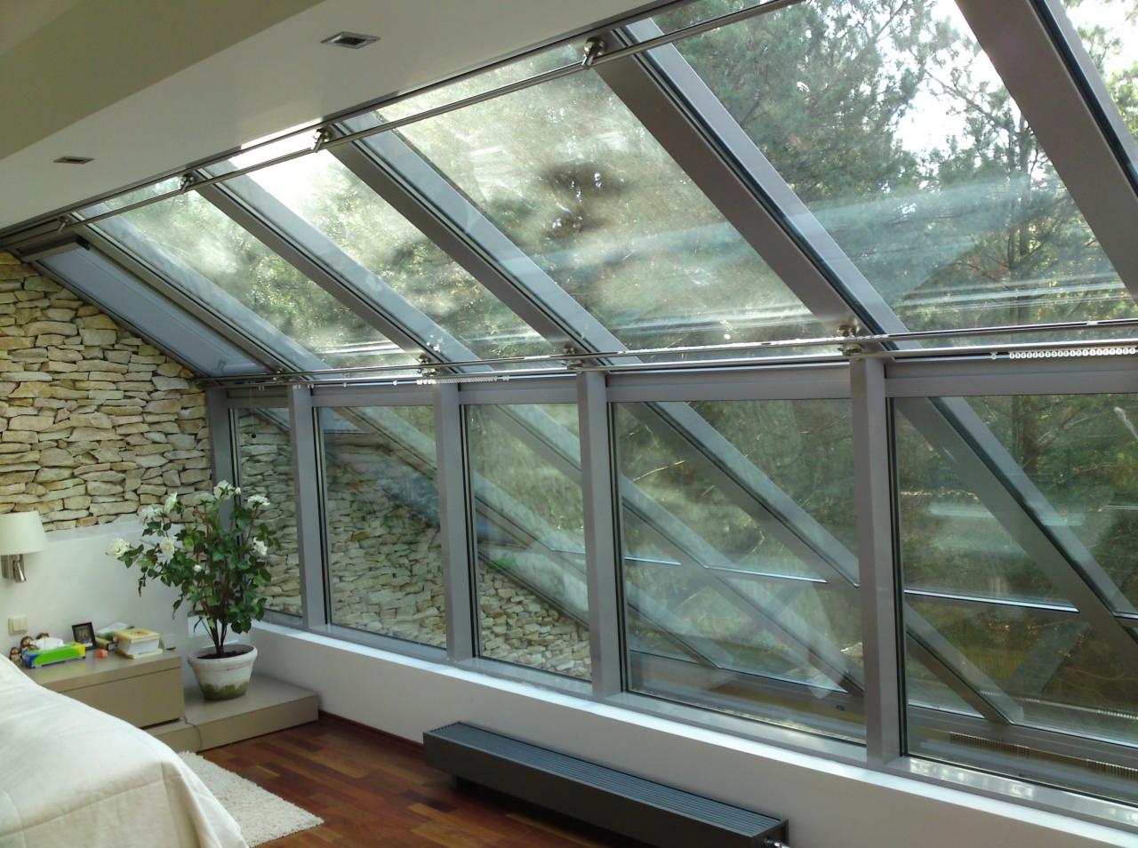 Nowoczesna oranżeria z przeszklonym dachem i ścianami, widok na zewnątrz, kamienna ściana w tle, drewniana podłoga, białe meble i roślina doniczkowa.