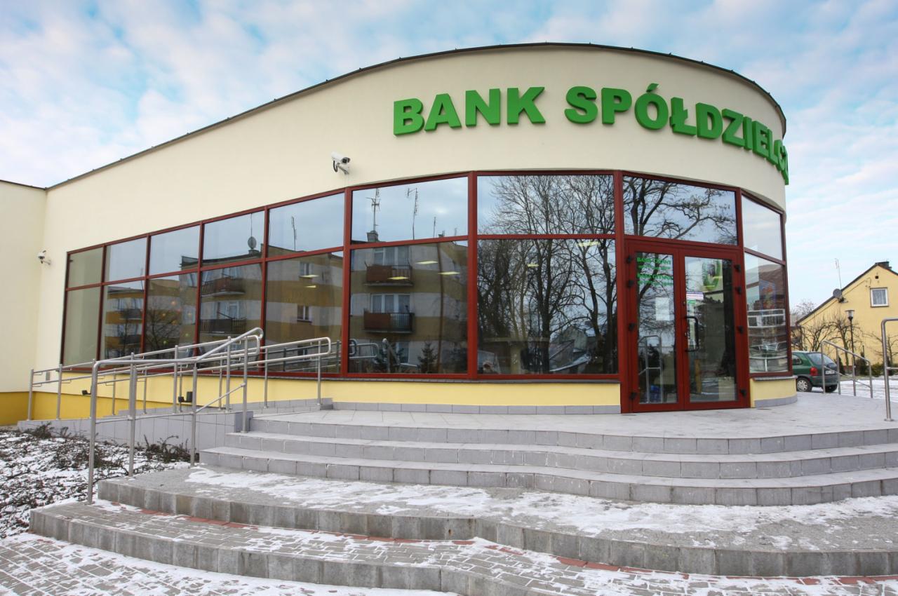 Budynek Banku Spółdzielczego z dużymi oknami i drzwiami wejściowymi, widoczne schody z podjazdem dla niepełnosprawnych, na zewnątrz lekki śnieg.