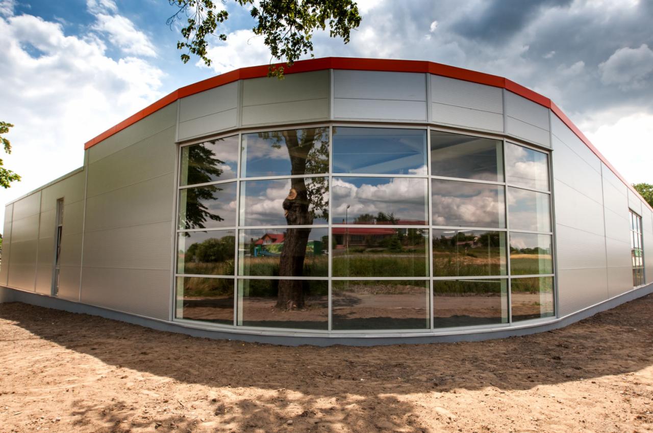 Nowoczesny budynek z panoramicznymi oknami z widocznym odbiciem drzewa i nieba, wykończony panelami w kolorze srebrnym z pomarańczowym akcentem na dachu.