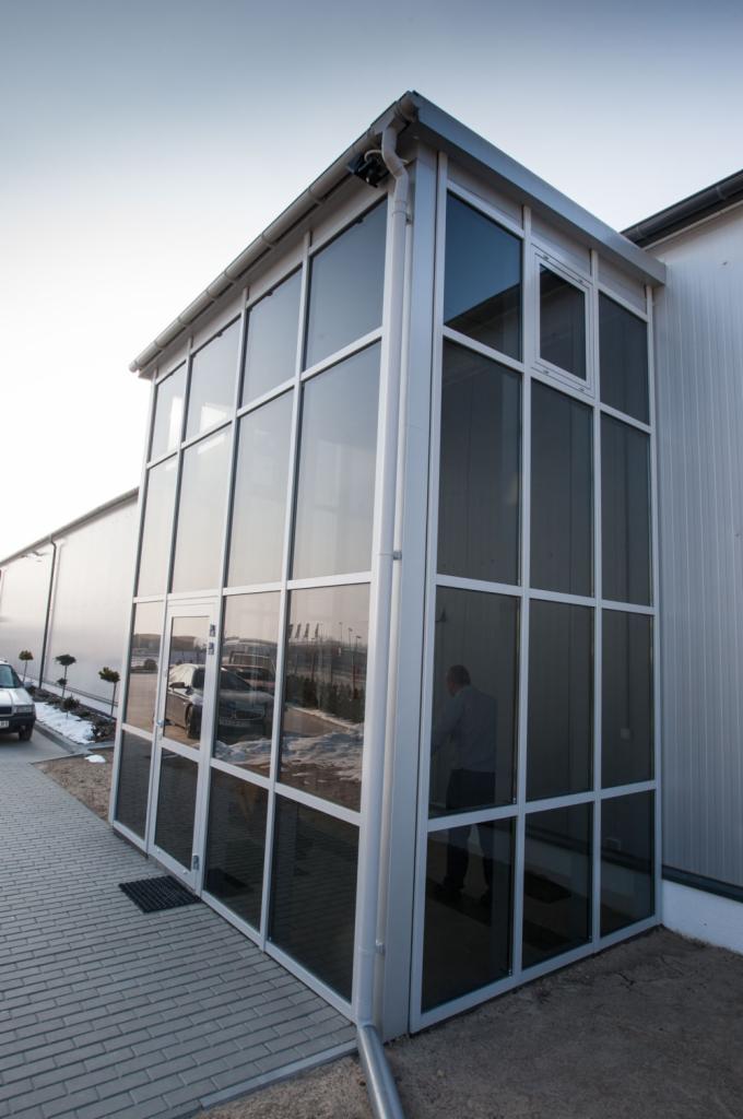 Szklana dobudówka do budynku przemysłowego z białymi ramami, widoczny fragment rynny, odbicie samochodów w szybach.