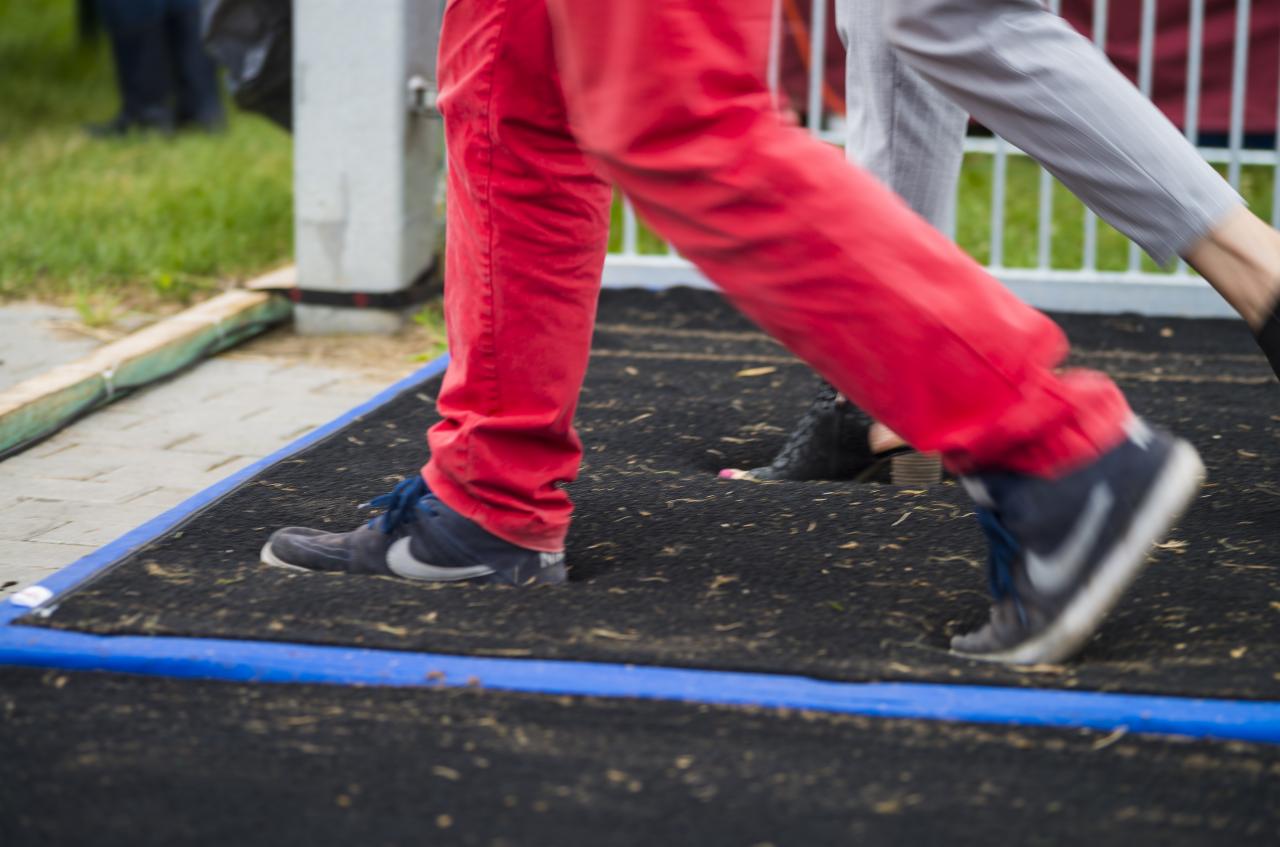 Osoby w ruchu na czarnej, gumowej nawierzchni z niebieską obwódką, prawdopodobnie plac zabaw lub teren rekreacyjny.