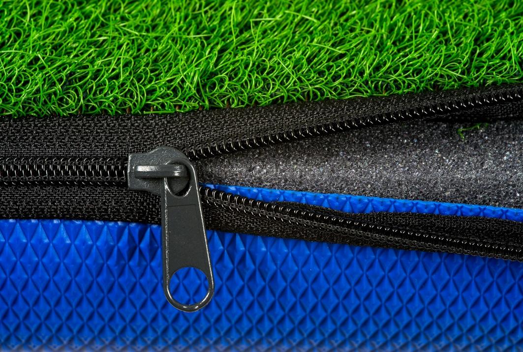 Warstwowa struktura podłoża: sztuczna trawa, czarny zamek błyskawiczny, szara pianka i niebieska struktura z wytłoczonym wzorem rombów.