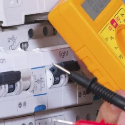 Pomiar napięcia na wyłączniku różnicowoprądowym z użyciem żółtego multimetru, widoczne przełączniki 'fan' i 'light'.