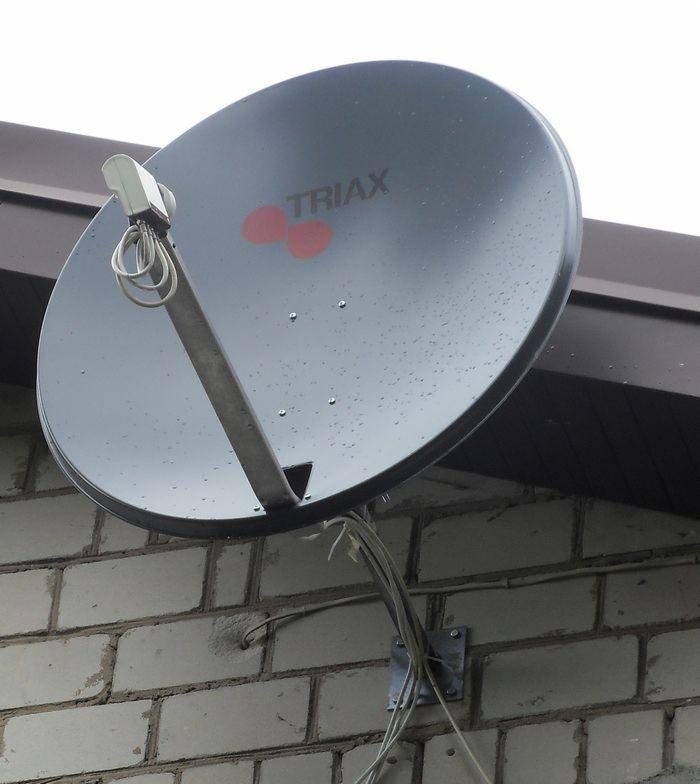 Szara antena satelitarna marki Triax zamontowana na ceglanej ścianie budynku, widoczne przewody antenowe zabezpieczone metalowym uchwytem.