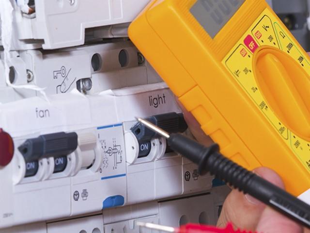 Pomiar napięcia na wyłączniku różnicowoprądowym z użyciem żółtego multimetru, widoczne przełączniki 'fan' i 'light'.