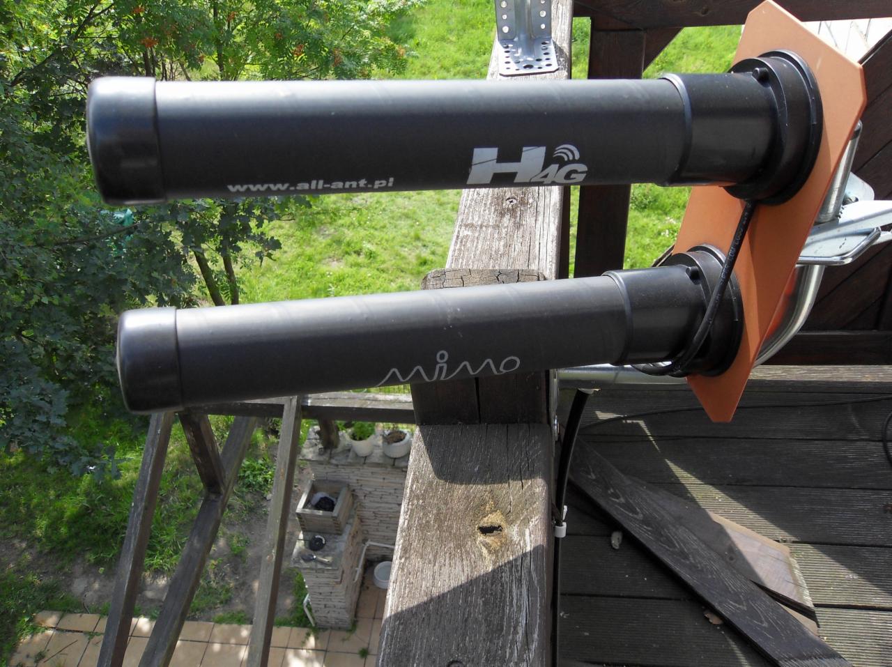 Dwie czarne anteny tubowe zamontowane na drewnianej konstrukcji z widocznymi napisami 'H4G' i 'mimo', tło z zielenią i fragmentem budynku.