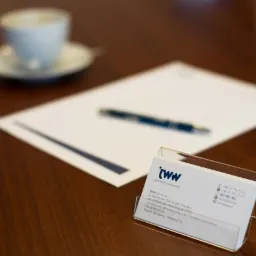 Wizytówka firmy księgowej 'JWW' w przezroczystym stojaku na biurku, w tle biała kartka papieru z długopisem i filiżanka kawy.