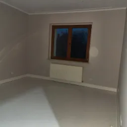 Pomalowane na szaro wnętrze pokoju z białymi listwami przypodłogowymi i oknem z drewnianą ramą, widok na ciemne drzewa za oknem.