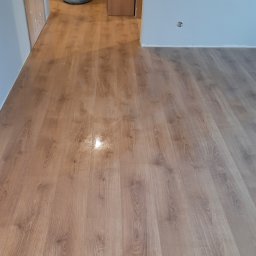 Piotr Gwiazda Usługi Remontowo-Wykończeniowe - Nowo ułożone panele podłogowe w jasnym odcieniu drewna, widoczne ślady po remoncie, takie jak wiadro i worek z odpadami w tle, oraz niedokończona szafka w przejściu.