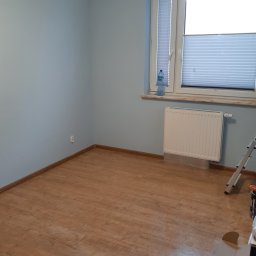 Piotr Gwiazda Usługi Remontowo-Wykończeniowe - Pomalowane na jasnoniebiesko wnętrze pokoju z panelami podłogowymi, białym grzejnikiem pod oknem i butelką wody na parapecie, widoczny fragment drabiny malarskiej.