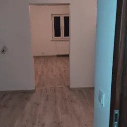 Pokój po remoncie z nową podłogą z paneli w odcieniach brązu, widok z korytarza na okno z grzejnikiem.