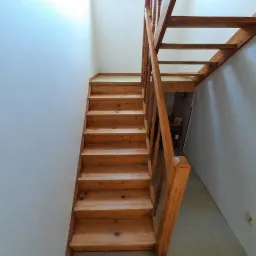 Drewniane schody z balustradą, widok z dołu, jasne drewno, białe ściany, widoczne słoje drewna.