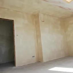 Wnętrze poddasza po nałożeniu gładzi, widoczne ściany i sufit w jasnym odcieniu beżu, surowy beton na podłodze, instalacja elektryczna wystająca ze ściany, otwór drzwiowy bez ościeżnicy.