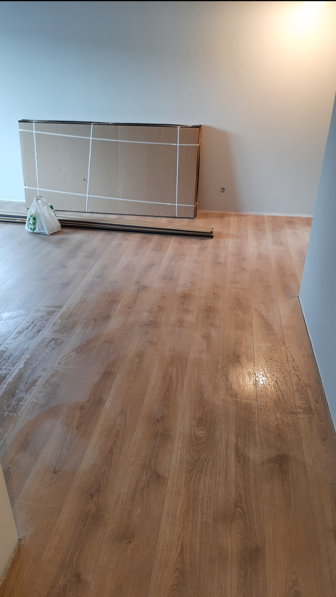 Przygotowanie do ułożenia paneli podłogowych: paczki paneli w kartonach oparte o ścianę, listwy przypodłogowe i torba foliowa na nowej, błyszczącej podłodze.