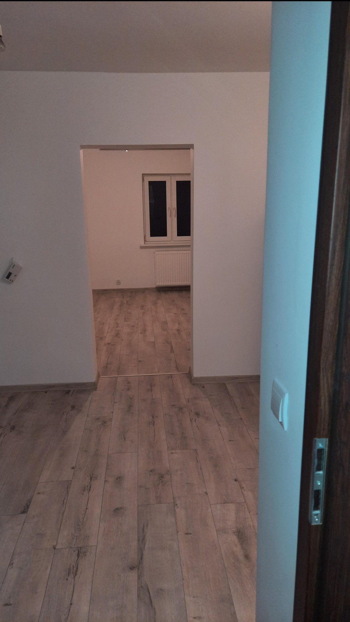 Pokój po remoncie z nową podłogą z paneli w odcieniach brązu, widok z korytarza na okno z grzejnikiem.