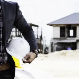 Mężczyzna w marynarce trzyma biały kask na tle budynku w trakcie budowy. Widoczny fragment planów. Kontrastowe światło podkreśla detale.