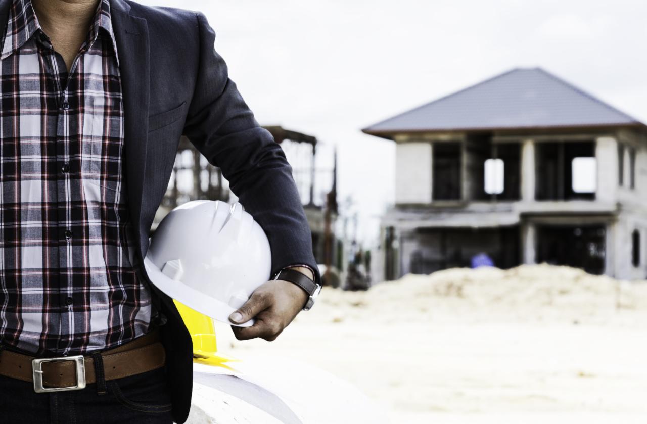 Mężczyzna w marynarce trzyma biały kask na tle budynku w trakcie budowy. Widoczny fragment planów. Kontrastowe światło podkreśla detale.