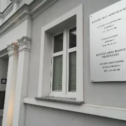 Elewacja budynku z oknem i tablicą informacyjną z nazwiskami adwokatów i radcy prawnego oraz numerami telefonów.