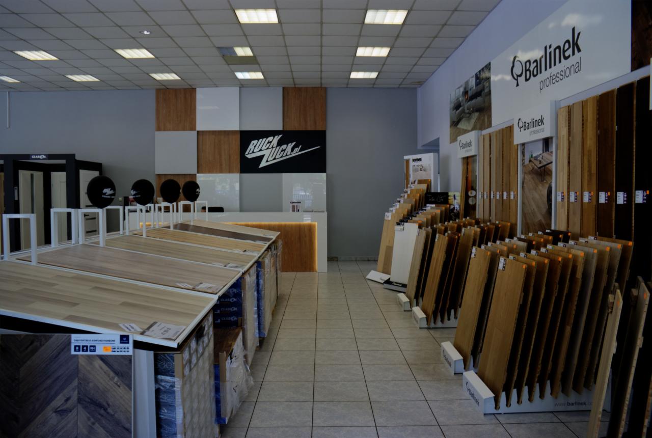 Ekspozycja różnorodnych paneli podłogowych w sklepie, widoczne próbki w różnych odcieniach drewna, logo producenta Barlinek na ścianie, w tle lada recepcyjna z logo RuckZuck.