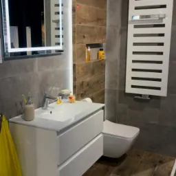 Nowoczesna łazienka z drewnopodobną podłogą, szarą glazurą, białymi meblami łazienkowymi, podświetlanym lustrem i grzejnikiem drabinkowym.