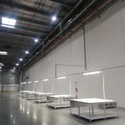 Wnętrze hali magazynowej z rzędem biurek pod linią świetlówek LED, widoczne instalacje pod sufitem i regały magazynowe w tle.