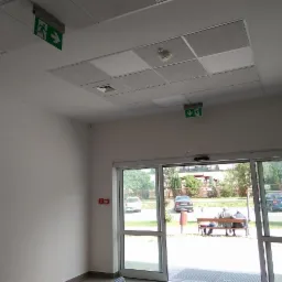 Wnętrze budynku z widocznymi znakami ewakuacyjnymi, czujnikiem dymu na suficie kasetonowym i automatycznymi drzwiami przesuwnymi prowadzącymi na zewnątrz.