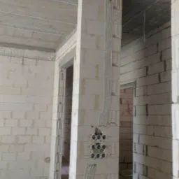 Surowy stan wnętrza budynku w trakcie prac elektrycznych: widoczne kable w peszlach prowadzone po ścianach z betonu komórkowego, puszki instalacyjne w ścianie, sufit betonowy z widocznymi przewodami.