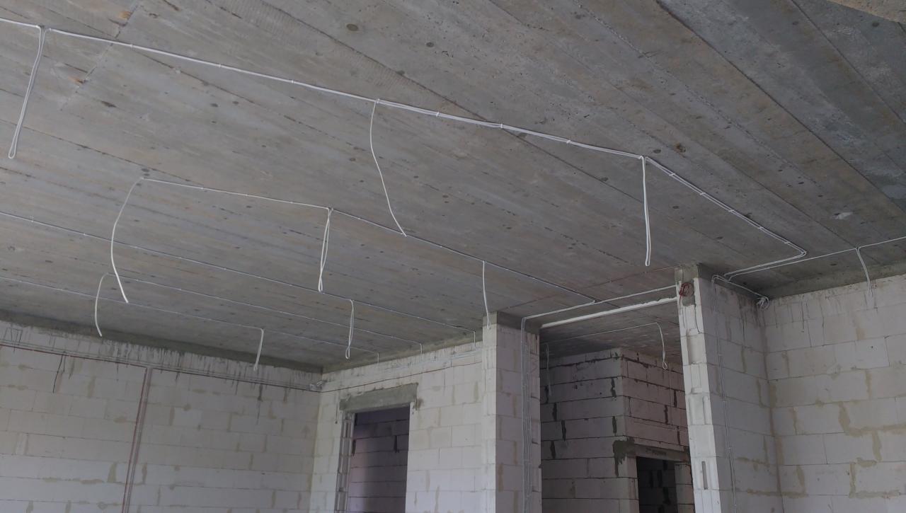Surowy stan wnętrza z widocznymi przewodami elektrycznymi prowadzonymi po betonowym suficie i ścianach z bloczków, przed tynkowaniem.
