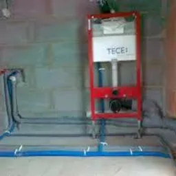 Stelaż podtynkowy WC marki TECE zamontowany na surowej ścianie z betonu komórkowego, z widocznymi niebieskimi rurami doprowadzającymi wodę, przymocowanymi do podłoża białymi uchwytami.