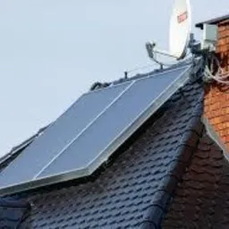 Panel solarny zamontowany na dachu pokrytym ciemną dachówką, obok ceglany komin z anteną satelitarną, widoczne przewody.