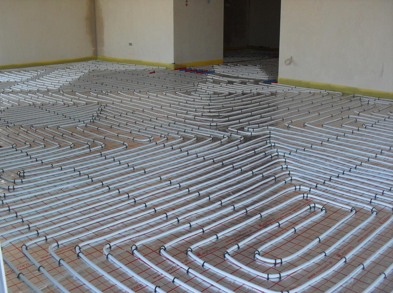 Białe rury systemu ogrzewania podłogowego rozłożone na siatce montażowej w surowym pomieszczeniu, widoczne żółte taśmy ochronne przy ścianach.