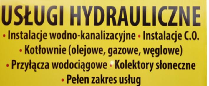 Żółta tablica reklamowa z czarnym napisem 'USŁUGI HYDRAULICZNE' i listą usług: instalacje wodno-kanalizacyjne, instalacje C.O., kotłownie (olejowe, gazowe, węglowe), przyłącza wodociągowe...