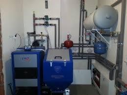 Złożona instalacja grzewcza w kotłowni z licznymi rurami, zbiornikami i urządzeniami sterującymi, w tym niebieska obudowa z panelem kontrolnym i szarą skrzynią z uchwytami.