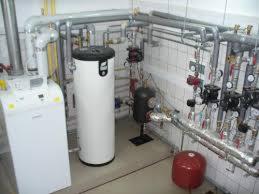 Kotłownia z białym zasobnikiem ciepłej wody, piecem gazowym, licznymi rurami i zaworami na tle białych płytek ściennych.