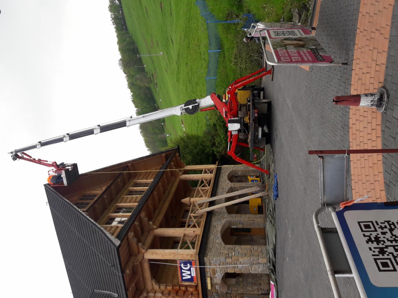 Pracownik w pomarańczowym kasku w koszu podnośnika teleskopowego obok drewnianego budynku z kamienną podmurówką i czarnym dachem, na tle zielonego zbocza.