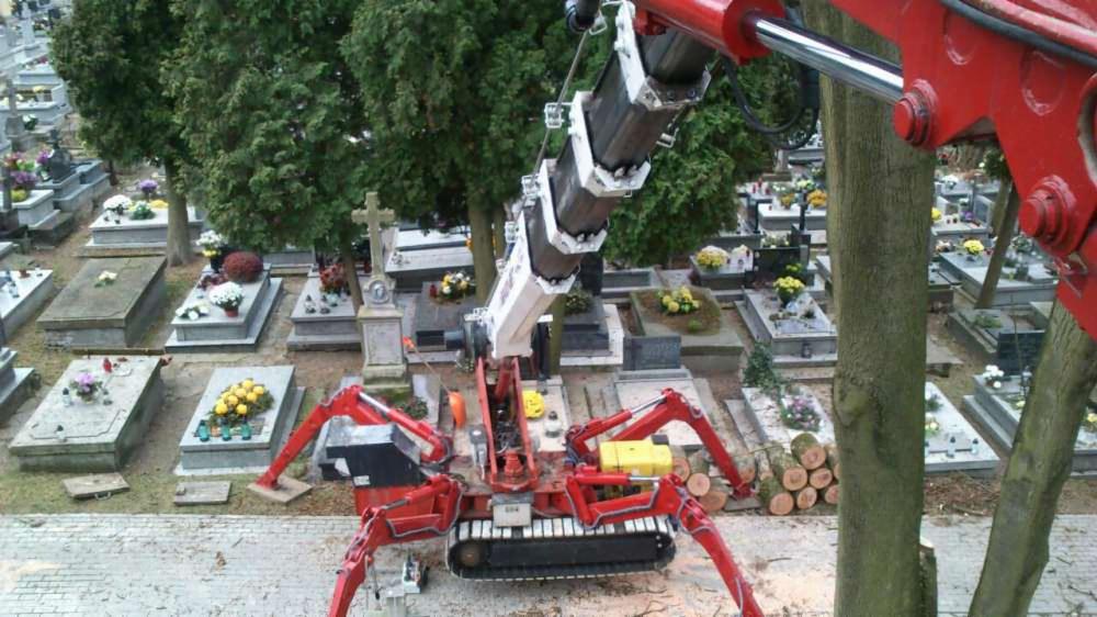 Czerwony dźwig pająk na gąsienicach, z wysięgnikiem nad drzewem, na tle cmentarza z nagrobkami i kwiatami.