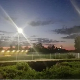 Rząd paneli słonecznych oświetlonych przez lampy uliczne wczesnym rankiem, z widocznym wschodzącym słońcem na horyzoncie.