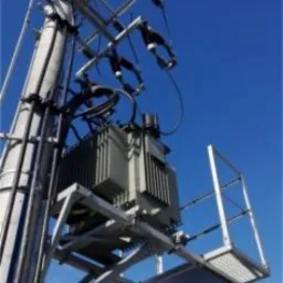 Transformator elektryczny zamontowany na słupie, widoczna platforma serwisowa, błękitne niebo w tle.