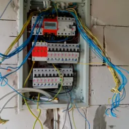 Rozdzielnica elektryczna w trakcie montażu w surowej ścianie z betonu komórkowego, widoczne liczne przewody o różnych kolorach, zabezpieczenia i wyłączniki.