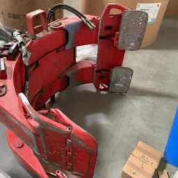 Czerwony, używany chwytak do wózka widłowego z hydraulicznymi przewodami, widoczne ślady użytkowania i otarcia farby, na tle betonowej podłogi magazynu.